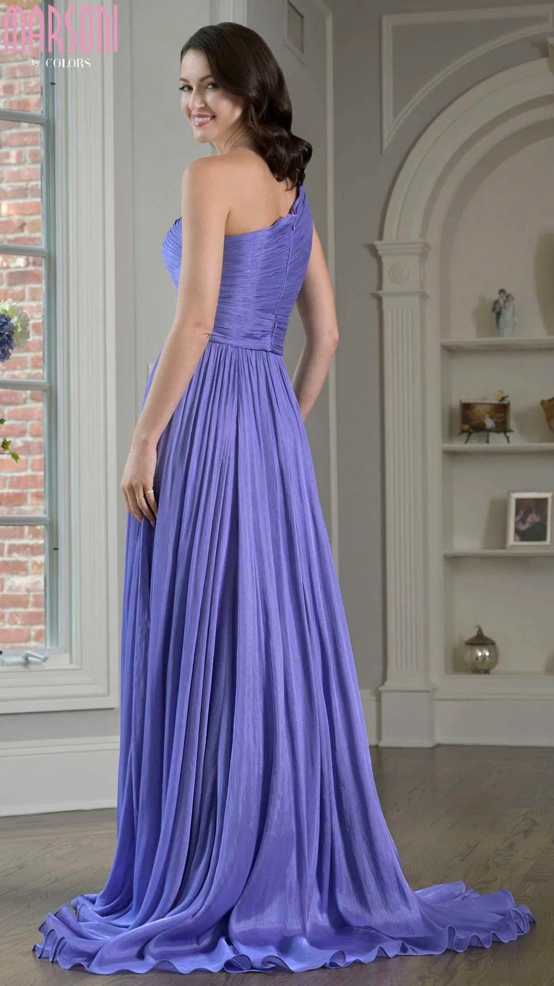 A woman stands indoors, smiling over her shoulder in the Marsoni M332—a floor-length, sleeveless purple chiffon formal gown.